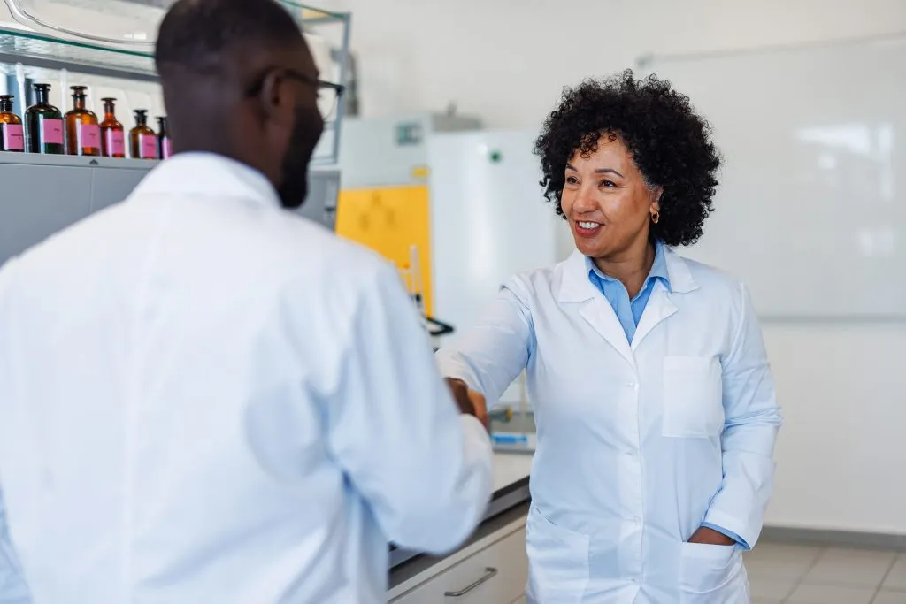 Two mature adult scientists wearing lab coats shaking hands in a laboratory, smiling and looking at each other, after a successful experiment